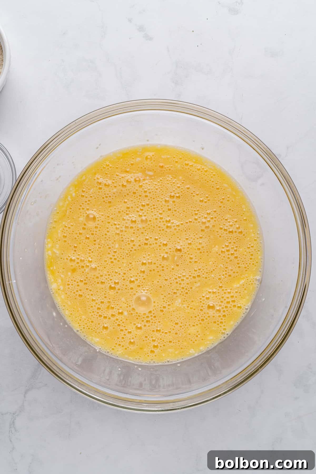 Power Breakfast Casserole 6 A glass bowl filled with beaten eggs, creating a frothy yellow mixture. The bowl rests on a light gray marbled surface.