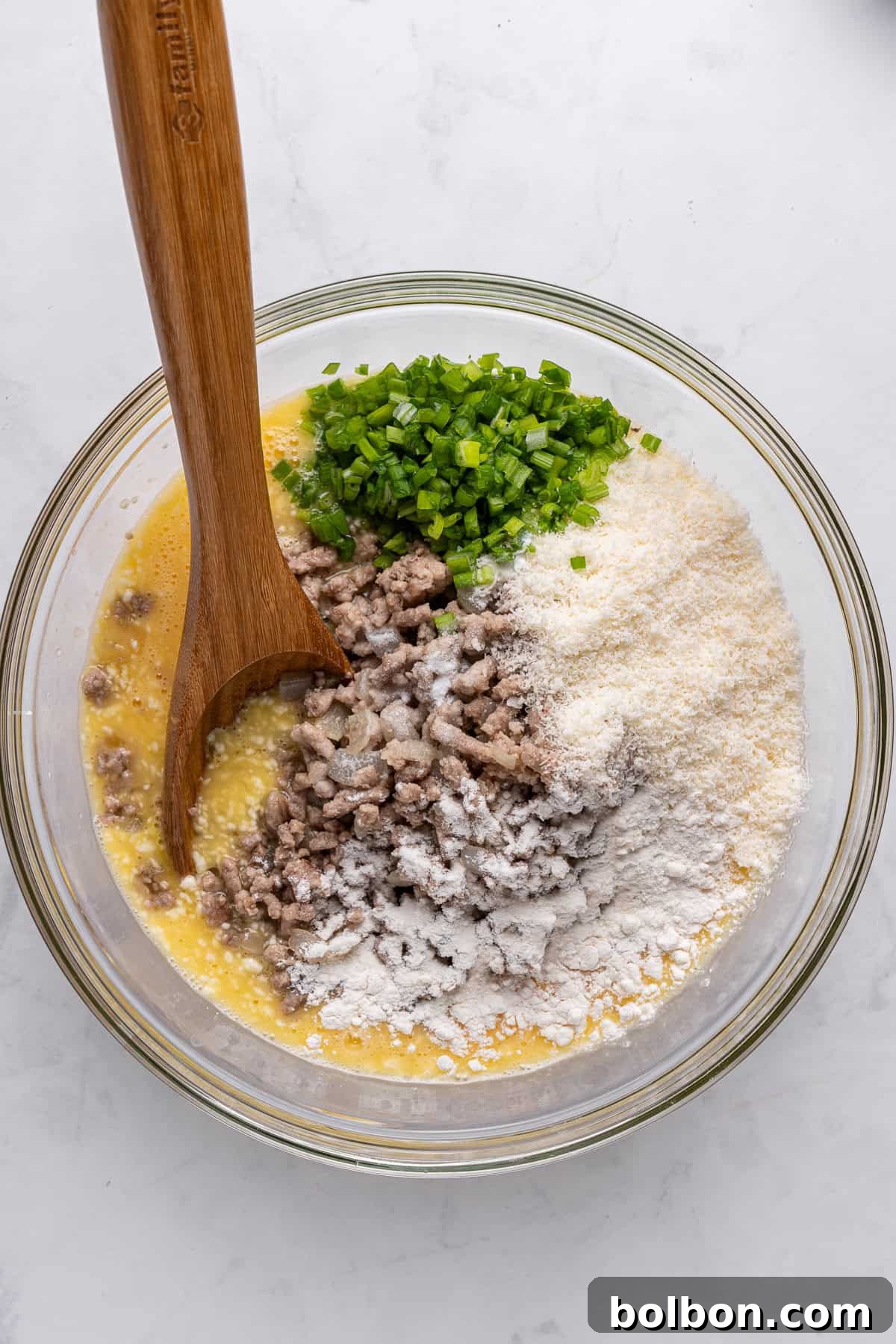 Power Breakfast Casserole 7 A glass bowl with grated cheese, chopped green onions, cooked sausage, and flour atop a mixture of beaten eggs. A wooden spoon is placed inside the bowl, ready to mix the ingredients. The bowl rests on a white marble surface.