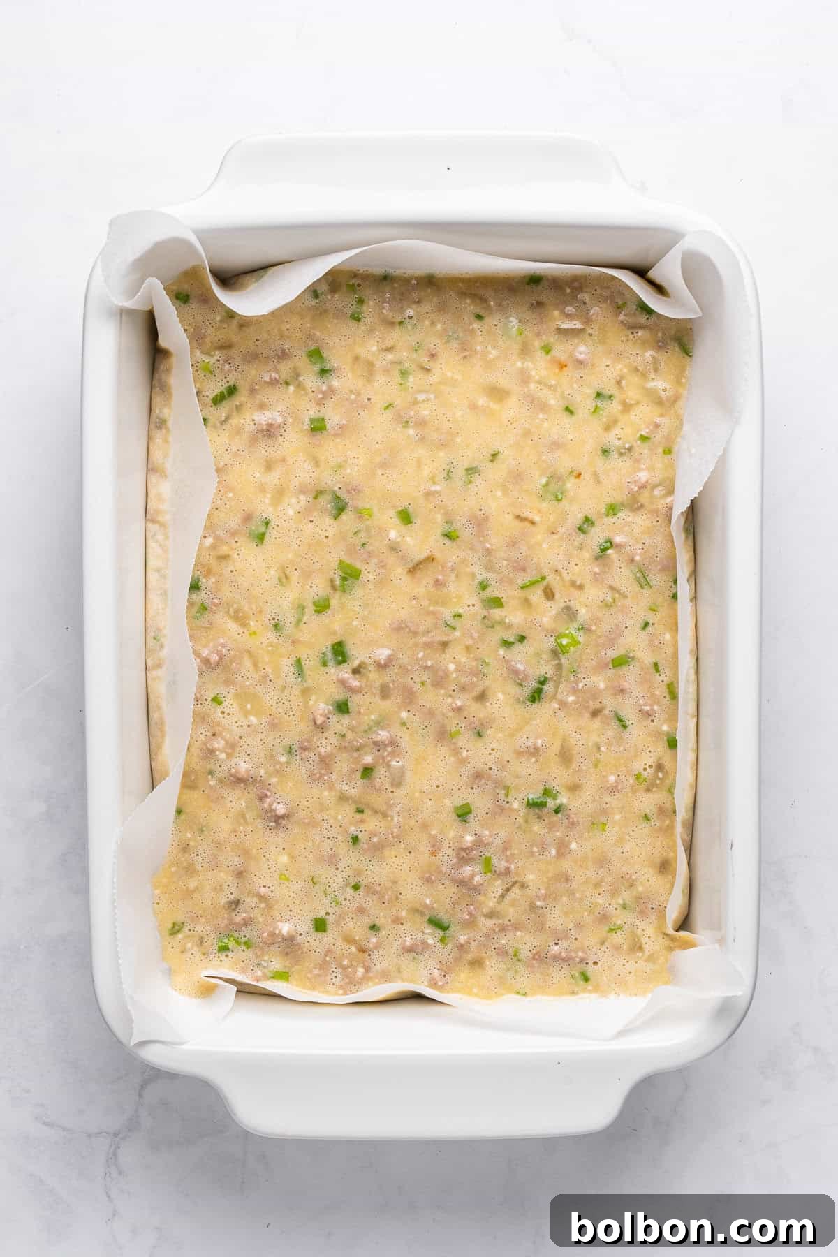 Power Breakfast Casserole 8 Overhead view of an uncooked egg and vegetable mixture in a parchment-lined white baking dish. The mixture includes visible green herbs and bits of vegetables.