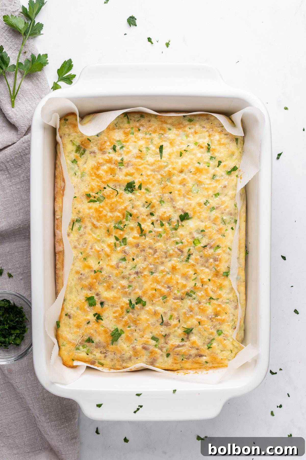 Power Breakfast Casserole 9 A freshly baked high protein breakfast casserole in a rectangular white dish. The casserole is topped with melted cheese and sprinkled with chopped herbs. A gray cloth and a small bowl of herbs are nearby on the white countertop.