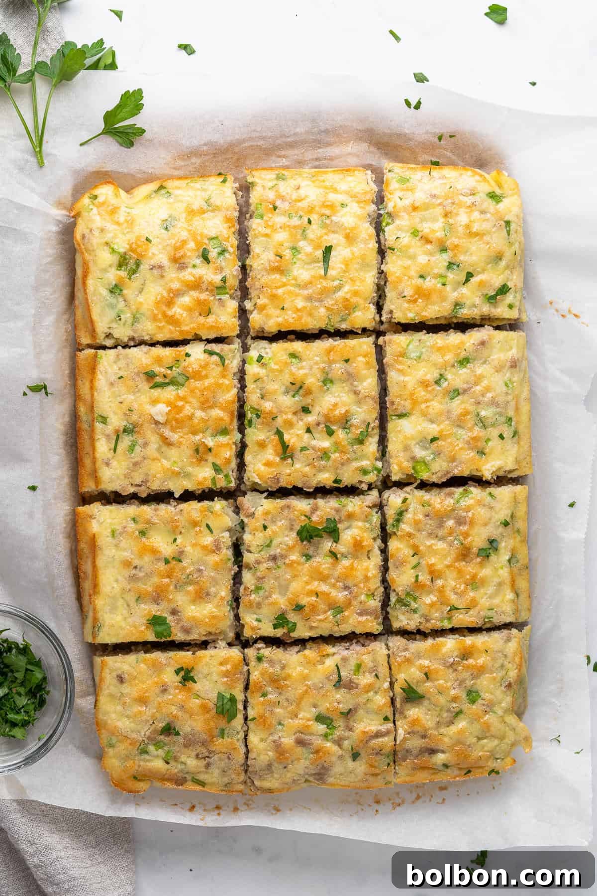 Power Breakfast Casserole 10 Golden, cheesy baked squares topped with chopped parsley on parchment paper. A small bowl of parsley is on the side, adding a touch of freshness to the presentation.