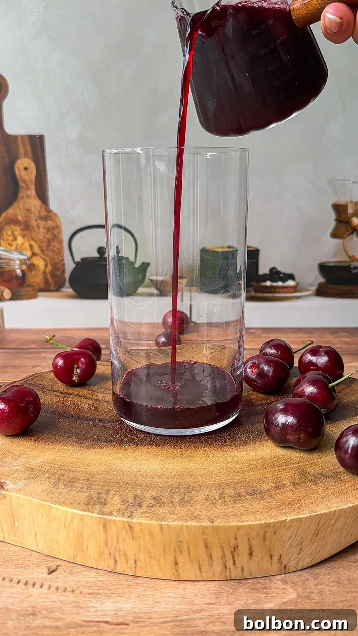 A hand pours dark red cherry juice from a glass pitcher into a clear glass on a wooden cutting board, surrounded by fresh cherries; a kitchen scene is visible in the background.