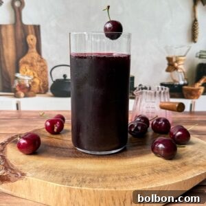 A glass of dark cherry juice sits on a wooden board, surrounded by fresh cherries. A single cherry garnishes the rim of the glass. The background shows a kitchen setting with utensils and cutting boards.