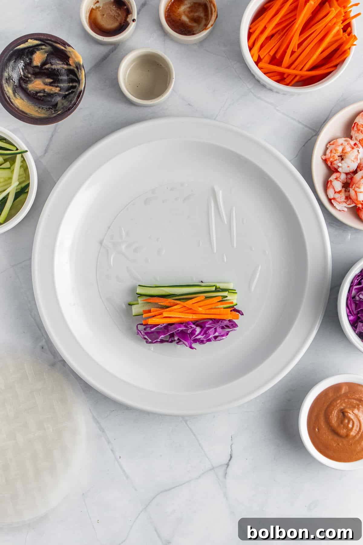 A plate with a rice paper sheet partially filled with julienned carrots, cucumbers, and purple cabbage, surrounded by bowls of shrimp, more vegetables, and various sauces, set on a light marble surface.