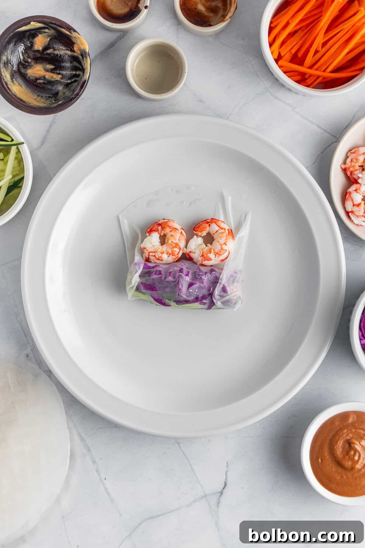 A partially assembled spring roll on a white plate, surrounded by bowls of ingredients like sliced carrots, cucumbers, shrimp, purple cabbage, and dipping sauces. A rice paper sheet is laid out next to the plate on a marble countertop.