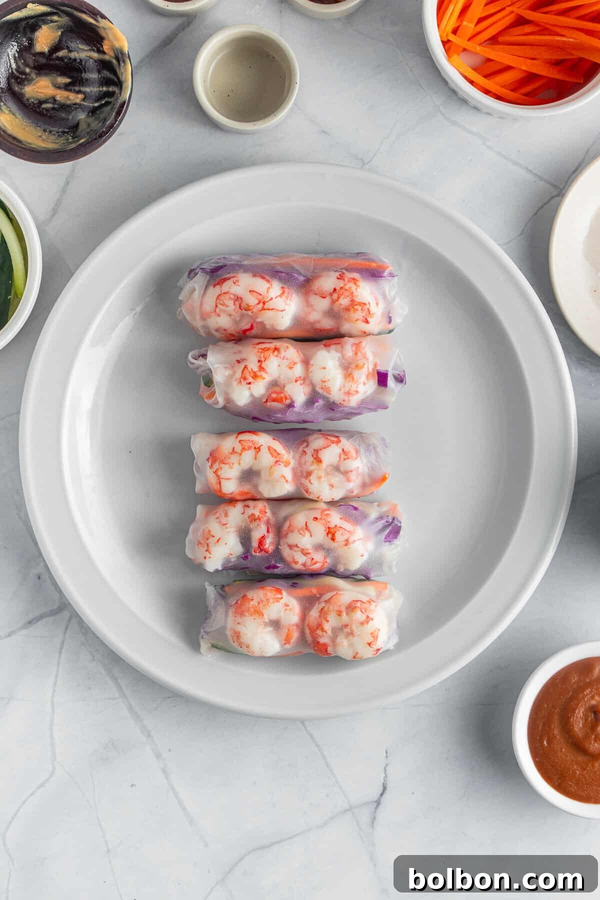 Four spring rolls are neatly arranged on a white plate. Surrounding the plate are small bowls containing various ingredients and sauces, including sliced vegetables and dipping sauces. The background is a marble surface.