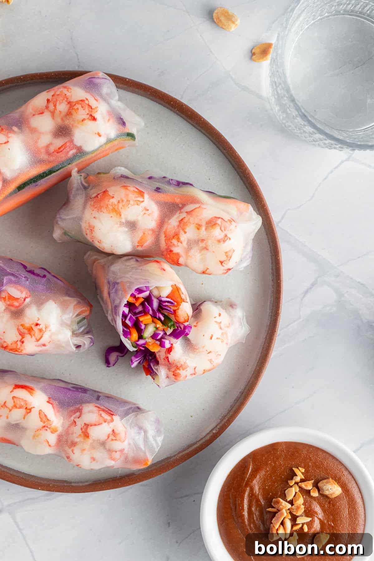 A plate of fresh spring rolls filled with shrimp, purple cabbage, and vegetables, partially unwrapped to reveal the colorful contents. Next to it, a bowl of peanut dipping sauce and a glass of water.