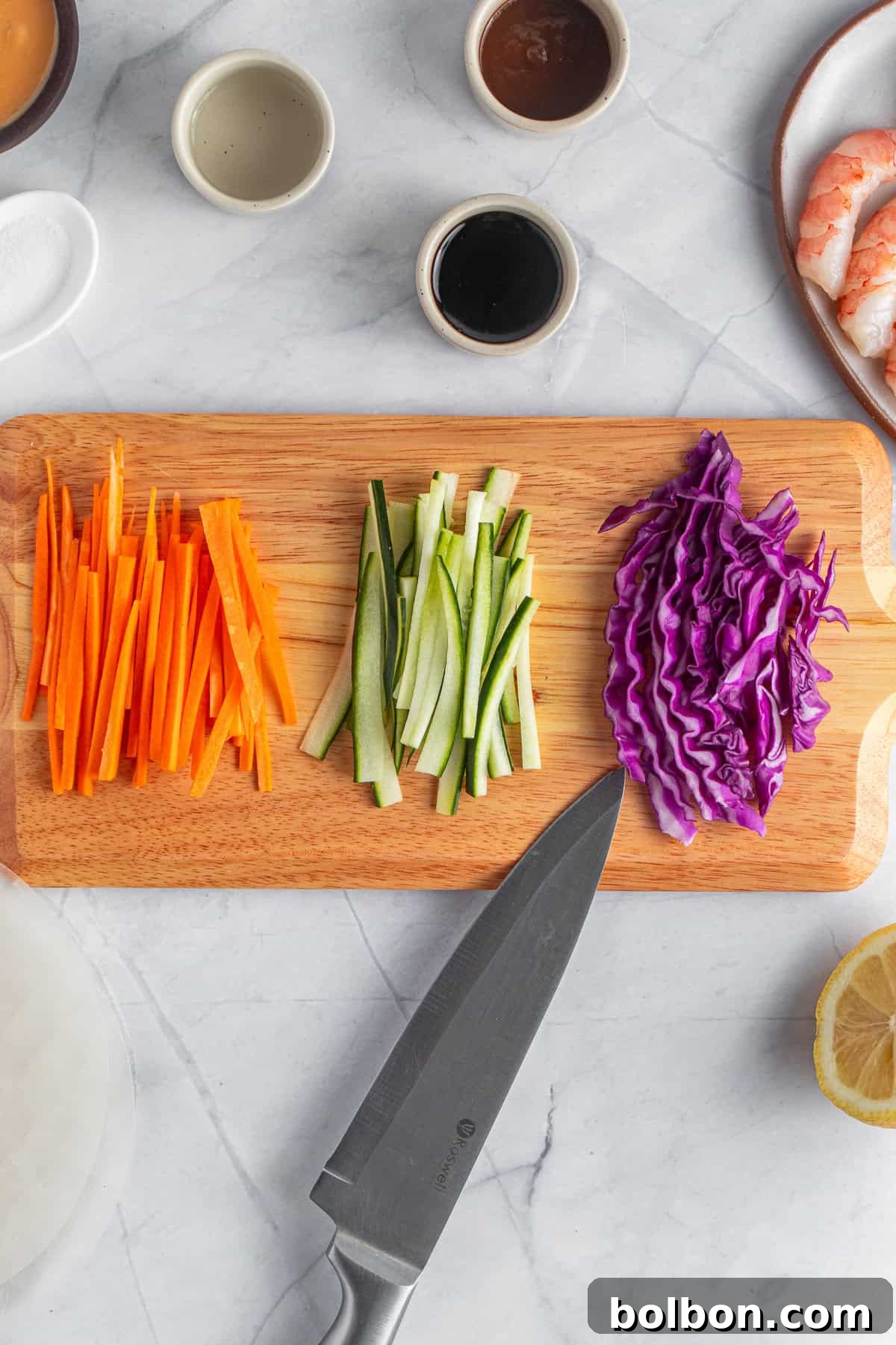 A wooden cutting board with sliced carrots, cucumbers, and shredded purple cabbage. A knife is placed nearby on the board. Surrounding the board are small bowls, shrimp, half a lemon, and a stack of rice paper.