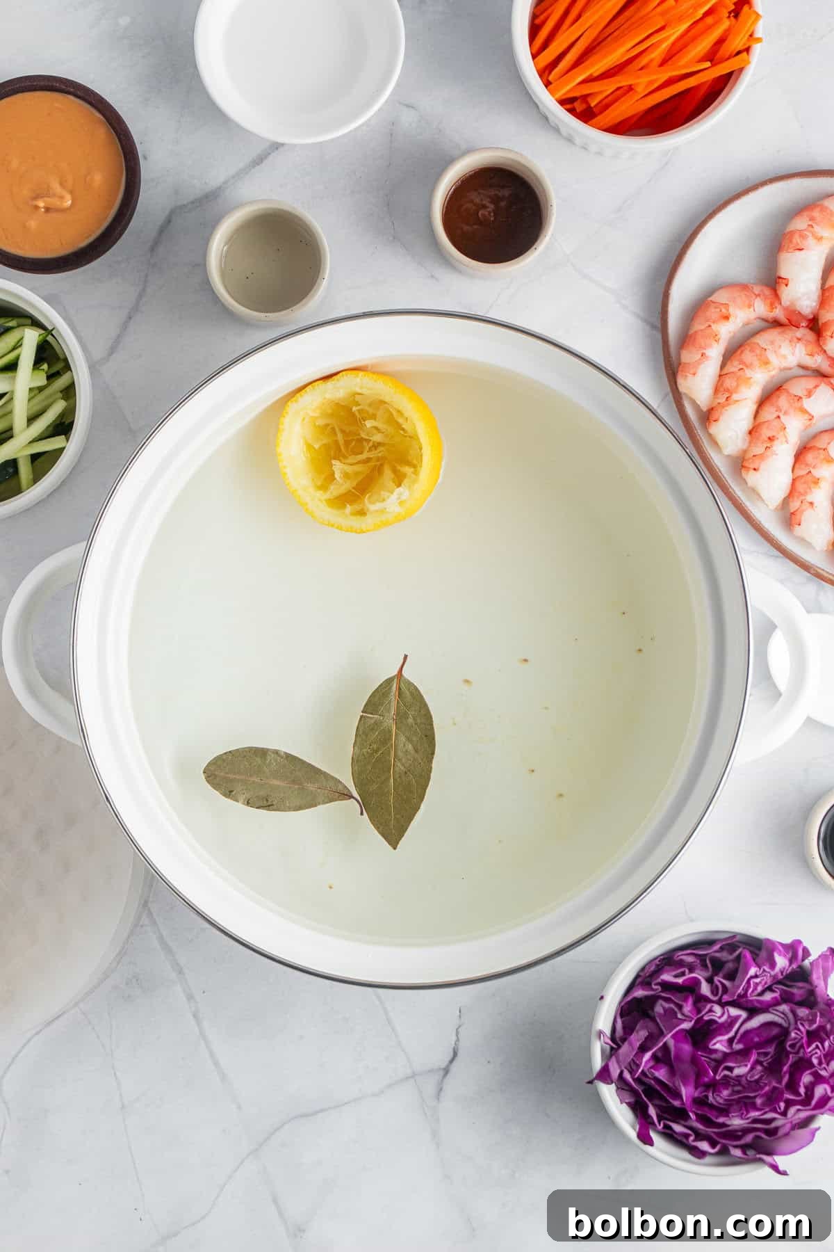 Ingredients for a dish are arranged around a white pot. The pot contains water with a lemon half and bay leaves. Surrounding the pot are shrimp, shredded purple cabbage, sliced carrots, and cucumber. Small bowls contain sauces and spices.
