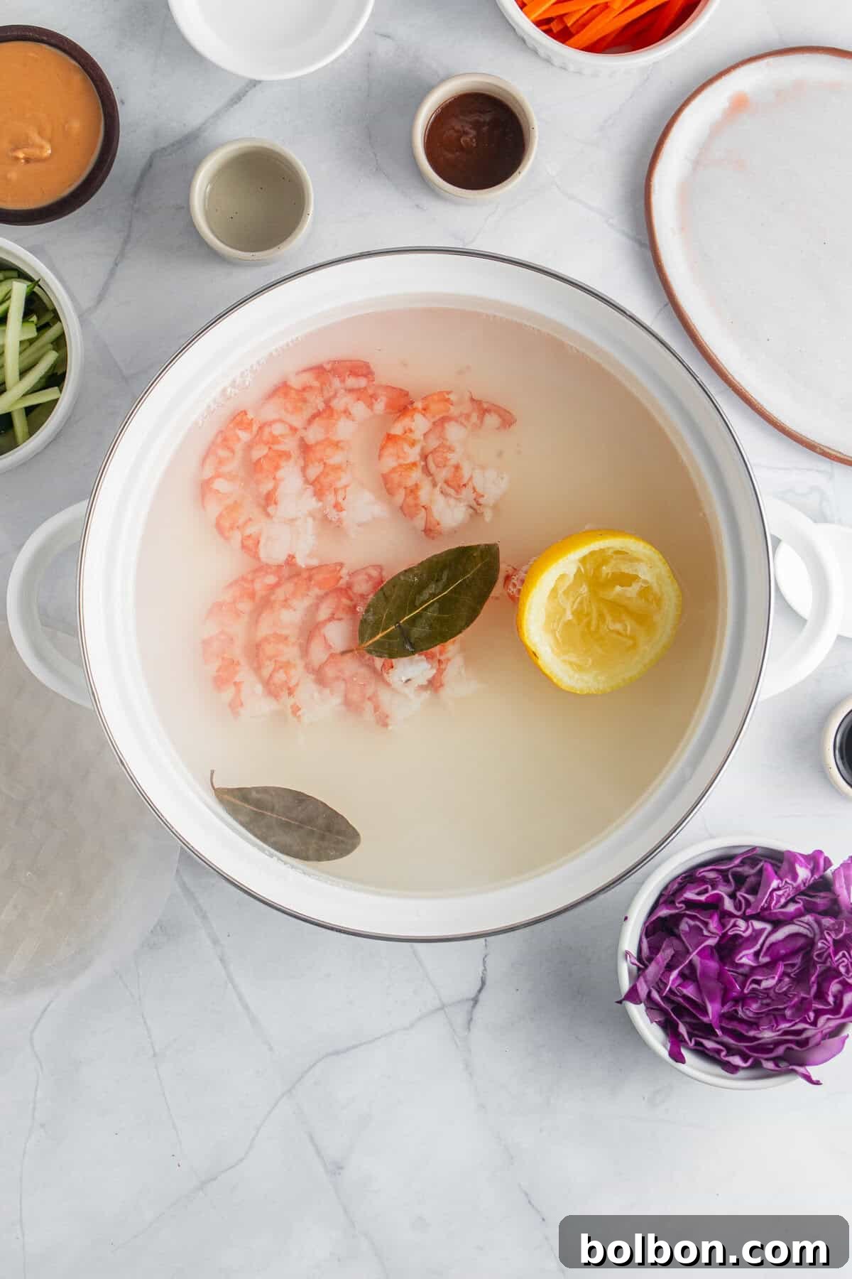 A pot filled with poaching water contains shrimp, a halved lemon, and bay leaves. Surrounding the pot are bowls with ingredients such as sliced vegetables, sauces, and spices on a marble countertop.