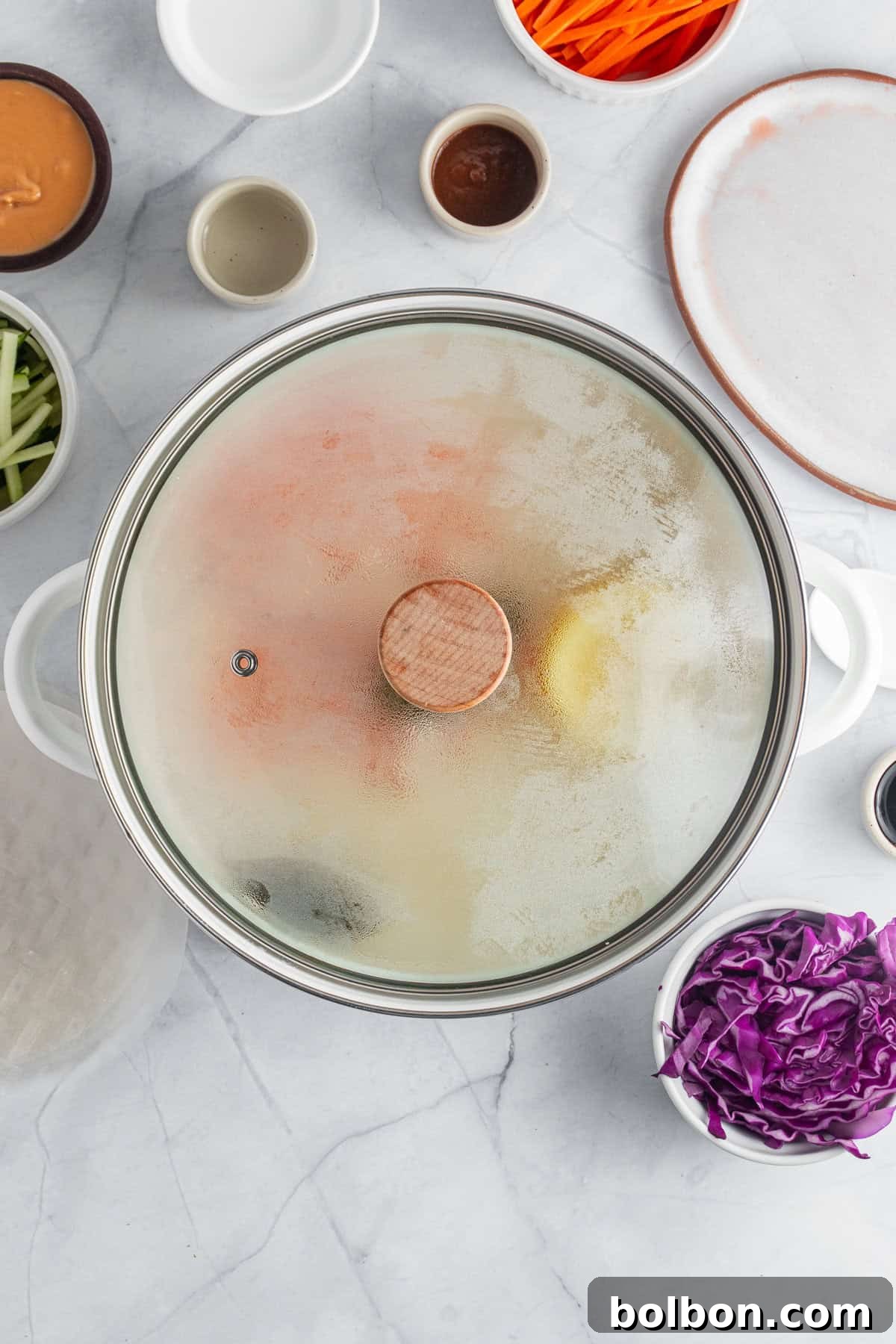 A pot with a glass lid is steaming on a marble countertop. Surrounding the pot are bowls of various ingredients, including sliced carrots, cucumbers, red cabbage, and small saucers of sauces, ready for cooking preparation.