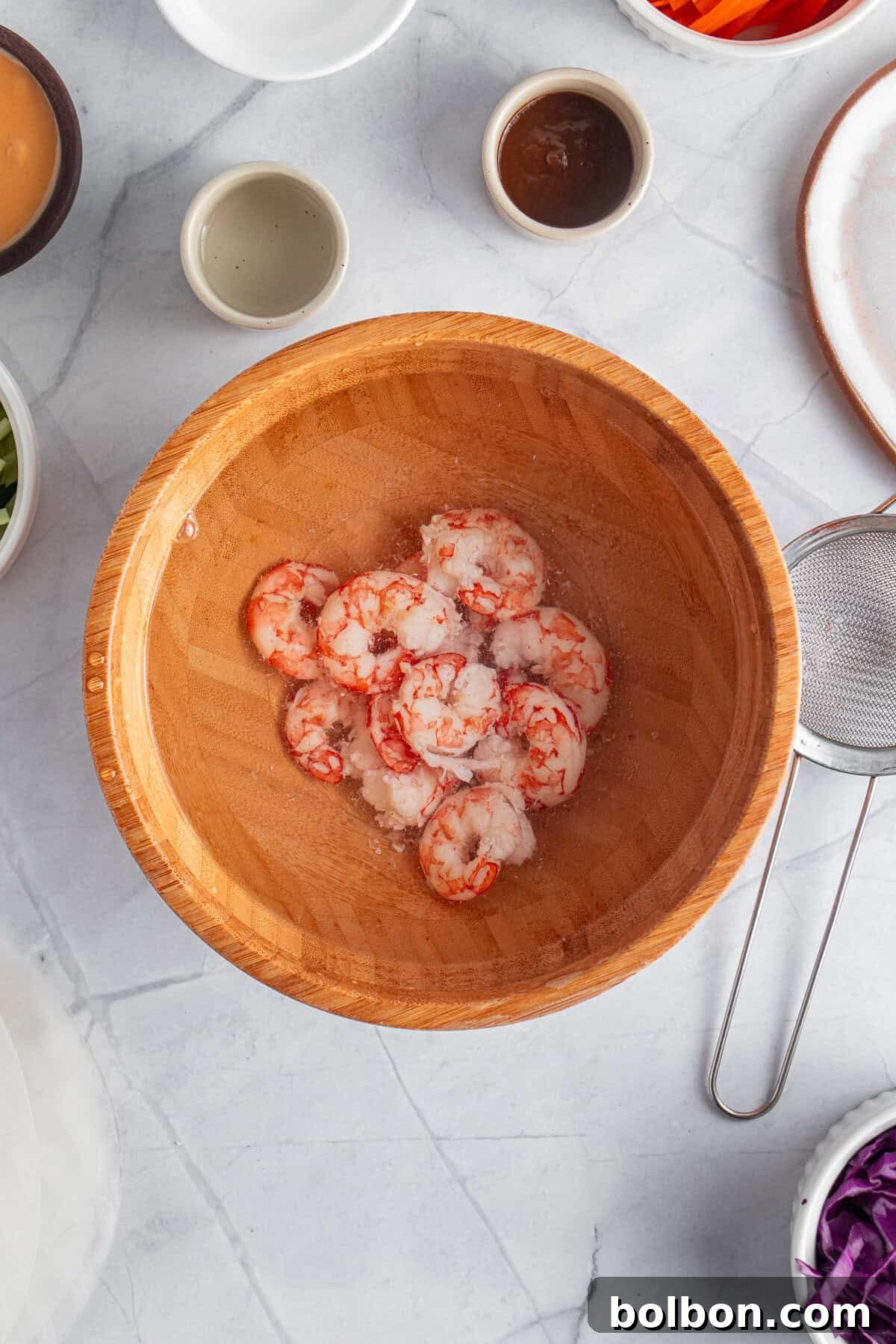A wooden bowl filled with shrimp sits on a marble countertop. Surrounding the bowl are small dishes with various ingredients and sauces, a strainer, and plates, suggesting preparations for a meal.