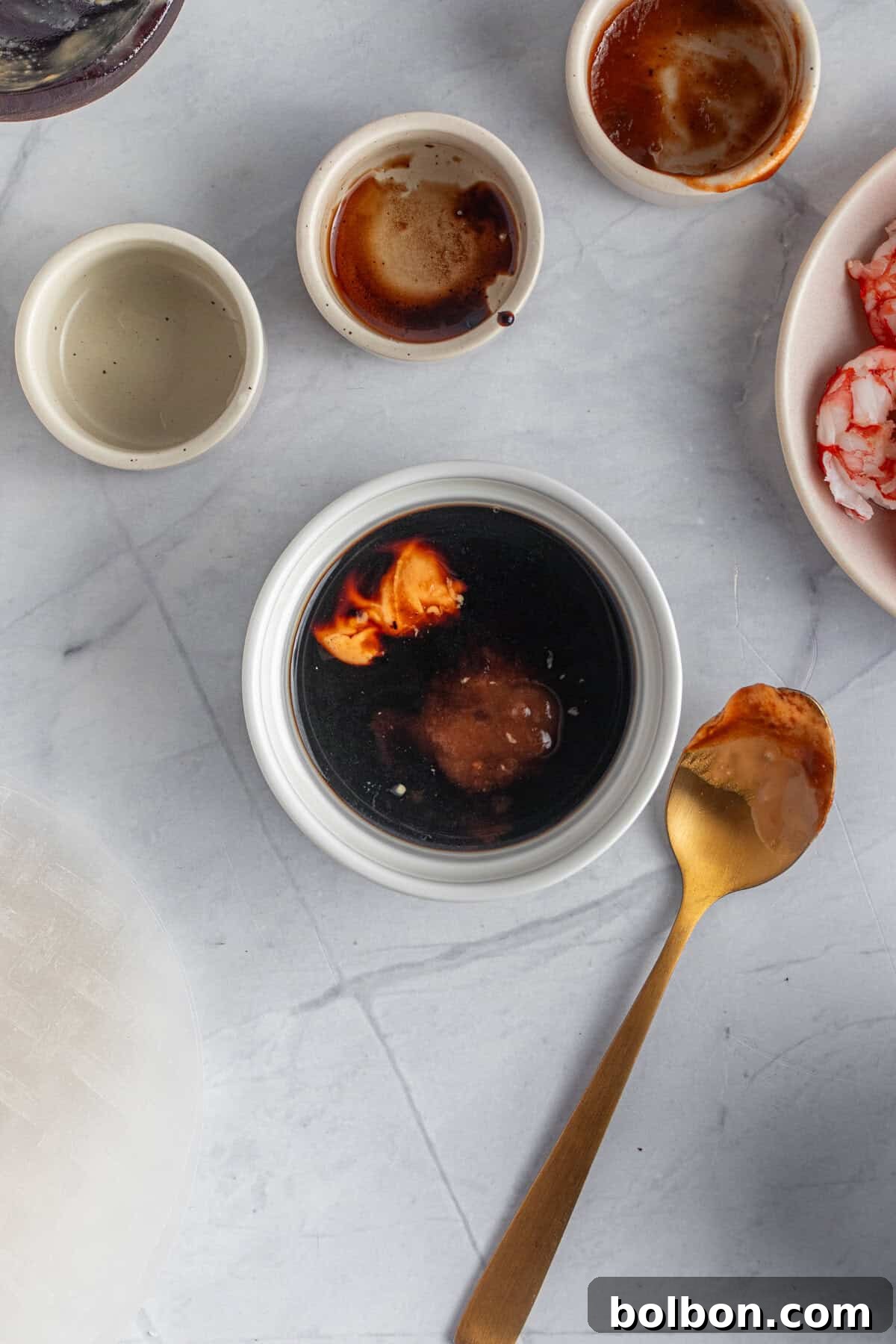 Various small bowls on a marble surface. One bowl contains a dark sauce with reddish garnish, and a golden spoon with sauce rests beside it. Other bowls hold remnants of ingredients and small bits of food, including shrimp.