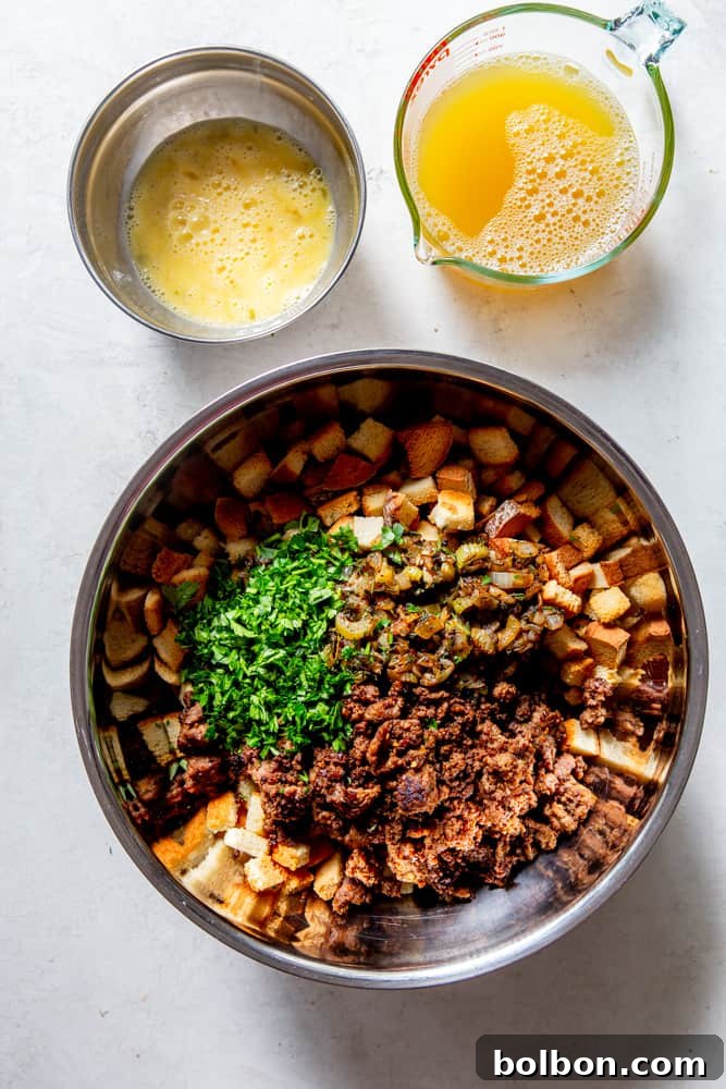 Timeless Savory Sausage Stuffing 9 Large steel bowl with toasted bread cubes, cooked sausage, minced parsley, and sautéed vegetables and herbs. Next to it are a measuring cup of chicken broth and a small bowl of beaten eggs.