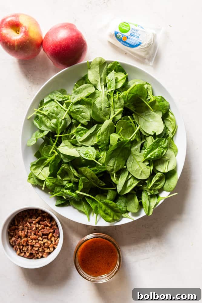 Ingredients for Apple Pecan Salad before assembling - a white bowl with baby spinach in it. Two whole red apples (Fuji), a small 4 ounce log of soft goats cheese, a small white bowl of chopped candied pecans, a jar of prepared apple cider vinaigrette.