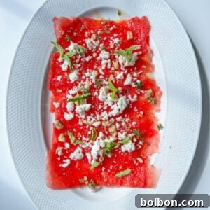 Watermelon Carpaccio: Thin slices of watermelon on an oval white plate, topped with crumbled white cheese, mint leaves, and a sprinkle of nuts or seeds, set against a white background.
