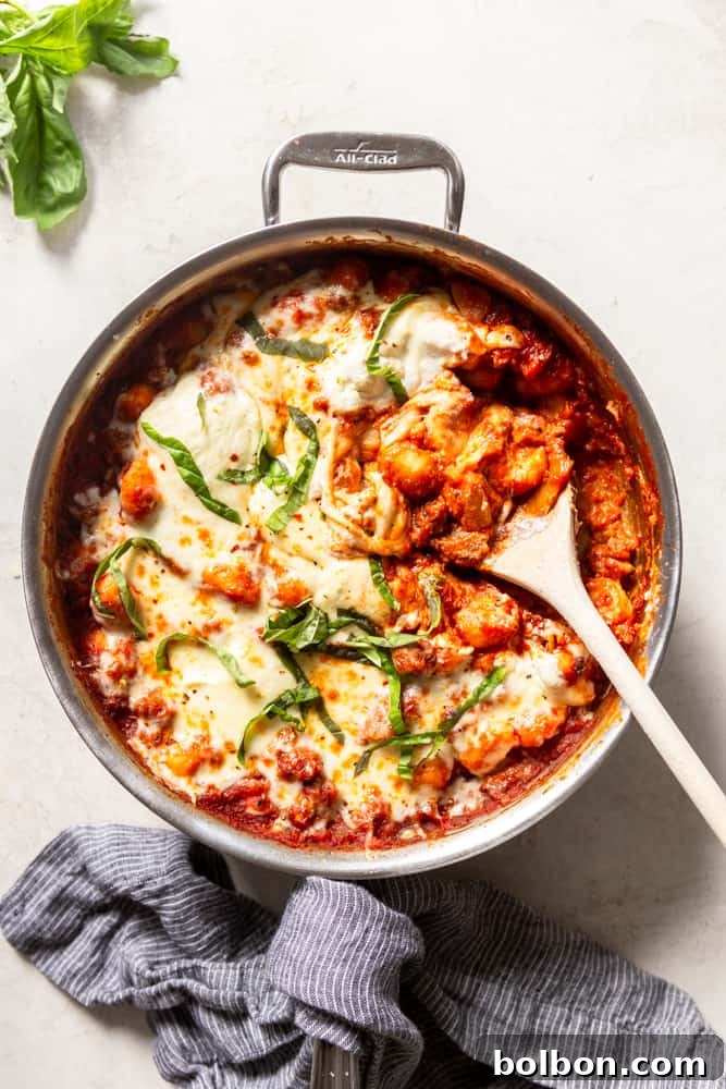 Hearty One-Pan Gnocchi & Sausage 2 Large stainless steel skillet with a vibrant red marinara sauce, tender gnocchi, and savory sausage, generously topped with melted mozzarella and dollops of creamy ricotta, garnished with fresh basil leaves.