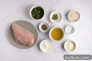 Effortless Weekly Chicken Bake 4 Raw chicken breast on a plate, surrounded by small bowls containing chopped spinach, pesto, rice, seasoning, broth, salt, mayonnaise, and grated cheese on a white marble surface.