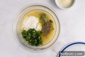 Effortless Weekly Chicken Bake 5 A glass bowl on a white surface containing yogurt, pesto, chopped greens, herbs, and a liquid mixture. A small bowl with grated cheese sits nearby.