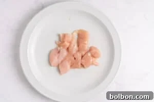 Effortless Weekly Chicken Bake 6 Raw, cubed pieces of chicken breast sprinkled with salt are arranged on a white plate, set against a light, marble-like background.