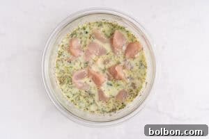 Effortless Weekly Chicken Bake 7 A glass bowl containing raw chicken pieces marinating in a creamy, herb-filled mixture, placed on a white marble surface.