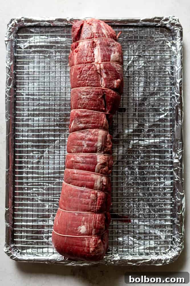 Uncooked Beef Tenderloin tied with kitchen twine, resting on a cooling rack over a foil-lined sheet pan, ready for seasoning.