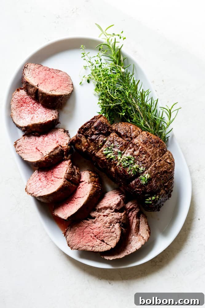 A glistening beef tenderloin roast, adorned with fresh green herbs, resting on a pristine white platter, ready to be served.