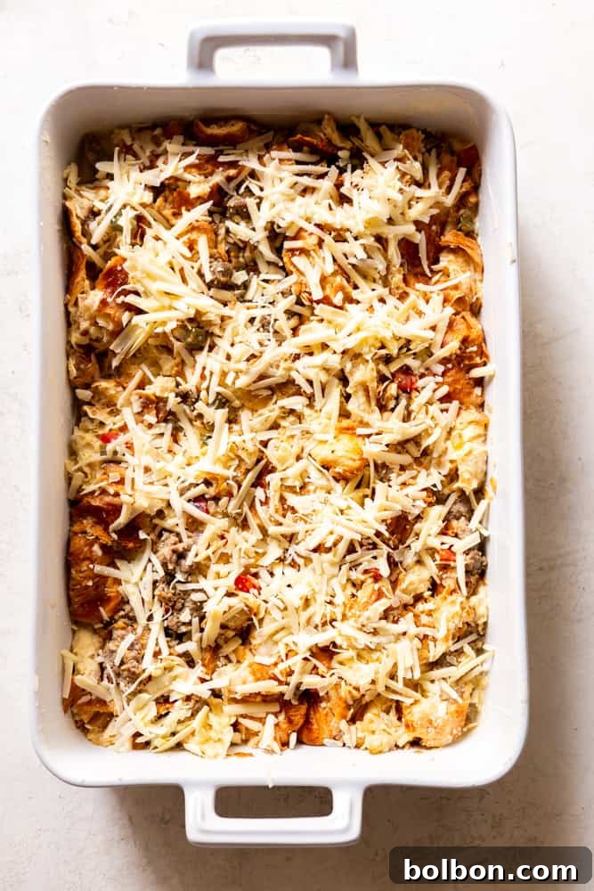 The Overnight Croissant Breakfast Casserole in a baking dish, topped with a generous layer of shredded cheese, ready to be baked.