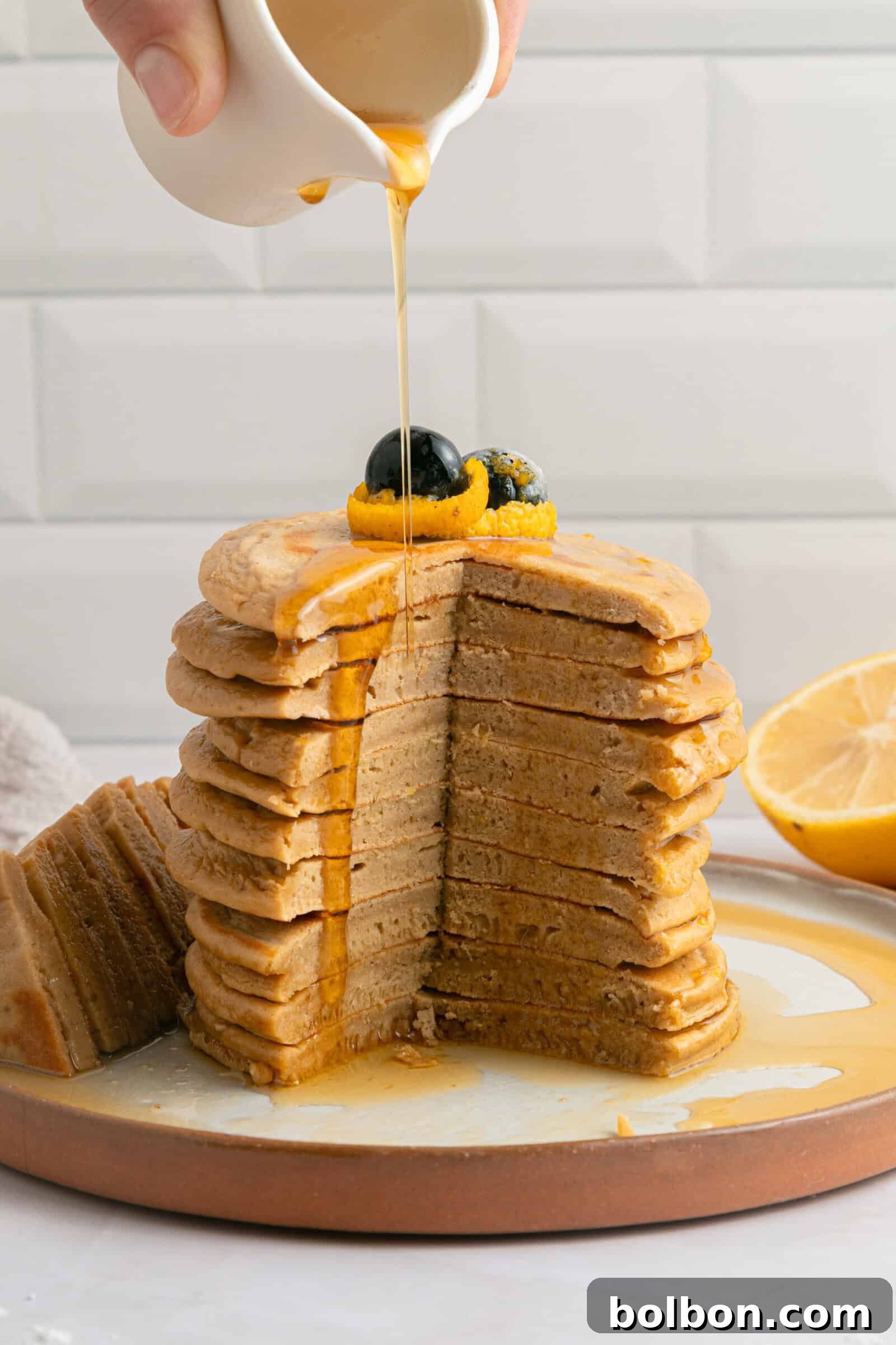 A stack of sliced pancakes topped with blueberries and an orange slice is drizzled with syrup from a small white pitcher, with a lemon half in the background.
