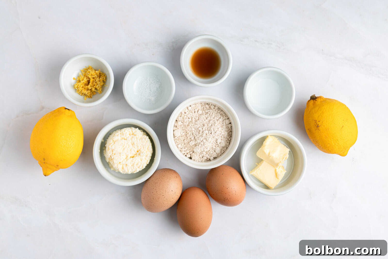A flat lay of baking ingredients on a white surface, including two whole lemons, three brown eggs, flour, butter, ricotta cheese, lemon zest, vanilla extract, baking powder, and two small bowls with clear liquids.