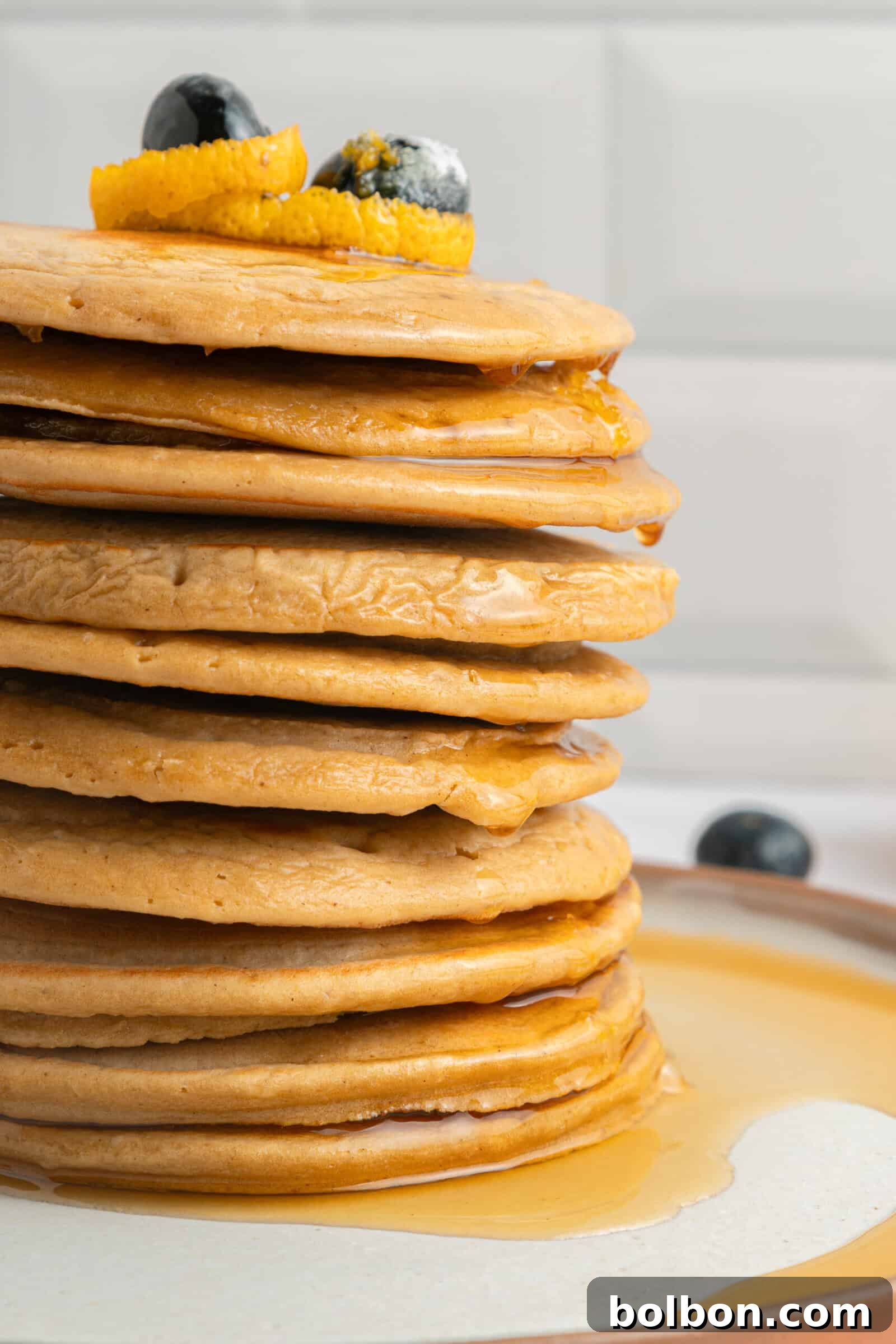 A tall stack of pancakes topped with orange peel and blueberries, with syrup dripping down the sides onto a plate. The background is a white tiled wall.