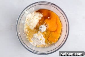 A food processor bowl containing eggs, flour, ricotta cheese, and other baking ingredients, viewed from above on a light countertop.