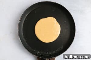 A pancake is cooking in a black nonstick frying pan, with batter spread in a round shape on the pan’s surface. The background is a light-colored countertop.