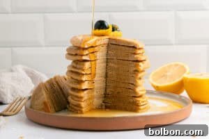 A tall stack of pancakes with a large wedge cut out is topped with blueberries and zest, with syrup being poured over. A halved lemon sits beside the plate on a white countertop.