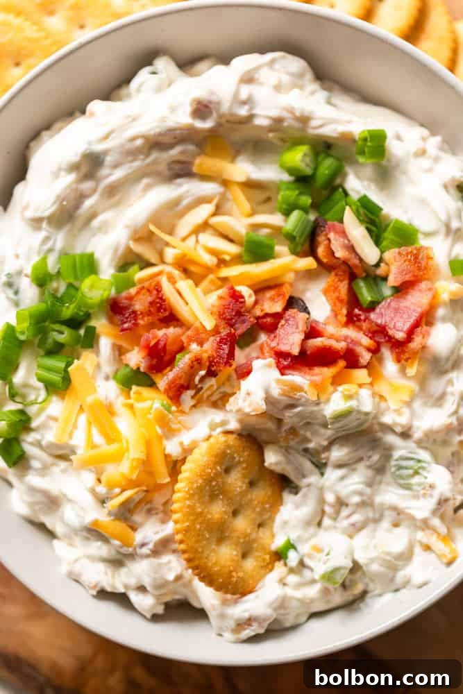 Irresistible Party Dip 2 Million Dollar Dip garnished with crispy bacon pieces, grated cheddar cheese, green onions, slivered almonds. A buttery cracker is being dipped.