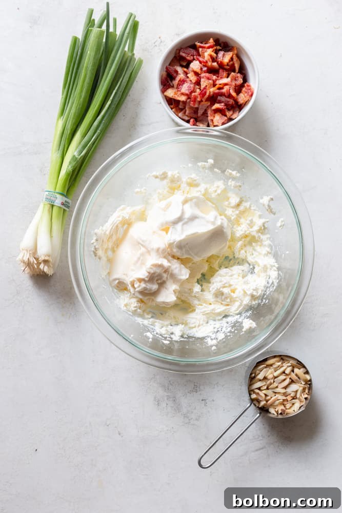 Irresistible Party Dip 4 Glass bowl with cream cheese, sour cream and may about to be mixed together. Surrounded by bacon, slivered almonds and green onions.