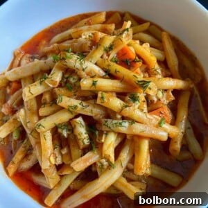 A white bowl filled with Fasolakia—braised green beans in a tomato-based sauce—garnished with herbs and small vegetable pieces, set against a dark background.