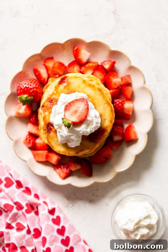 Strawberry pancakes topped with whipped cream and a fresh strawberry, on a scalloped plate, next to a bowl of whipped cream and a napkin with pink and red hearts.