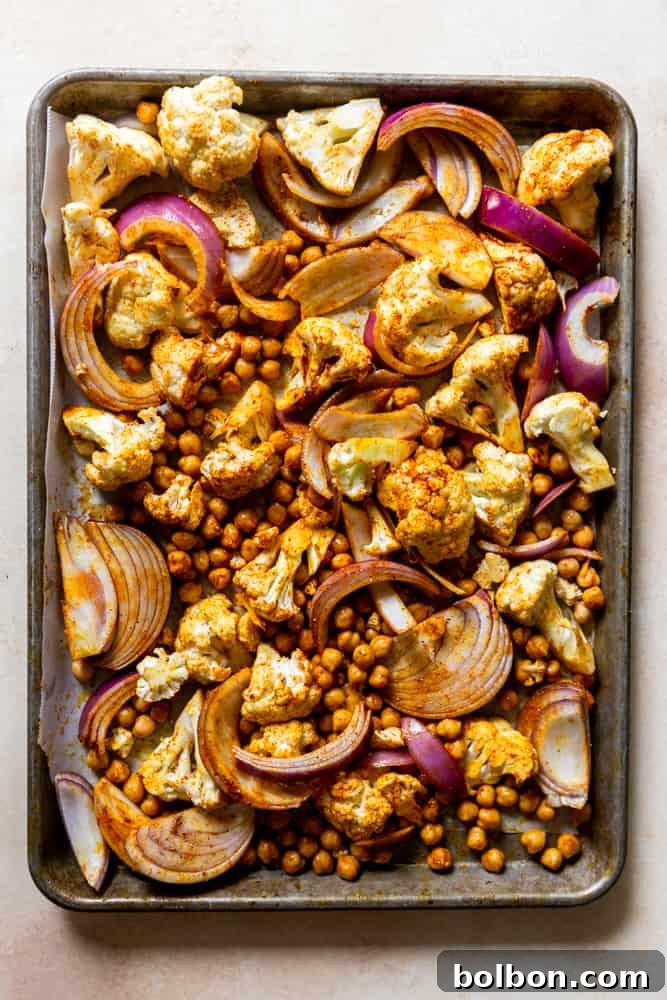 Sheet pan loaded with raw cauliflower florets, canned chickpeas, and sliced red onion, all generously coated in a vibrant blend of Shawarma-inspired spices and olive oil, ready for roasting.