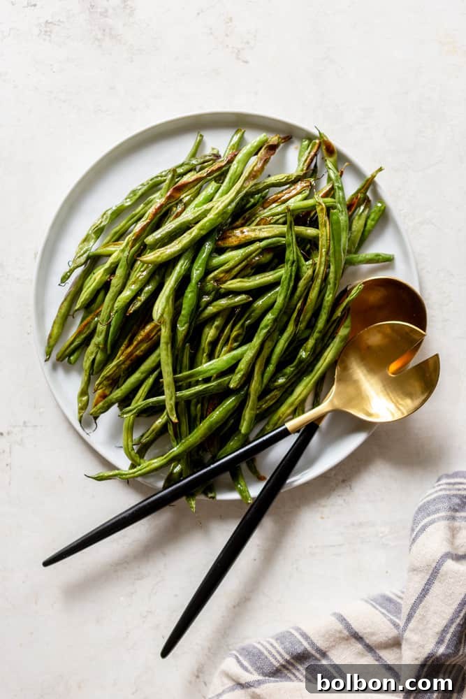 Perfectly roasted green beans served on a white plate with gold and black utensils. The setting includes a white background with a grey and blue striped dish towel nearby, emphasizing freshness and an inviting dining experience.