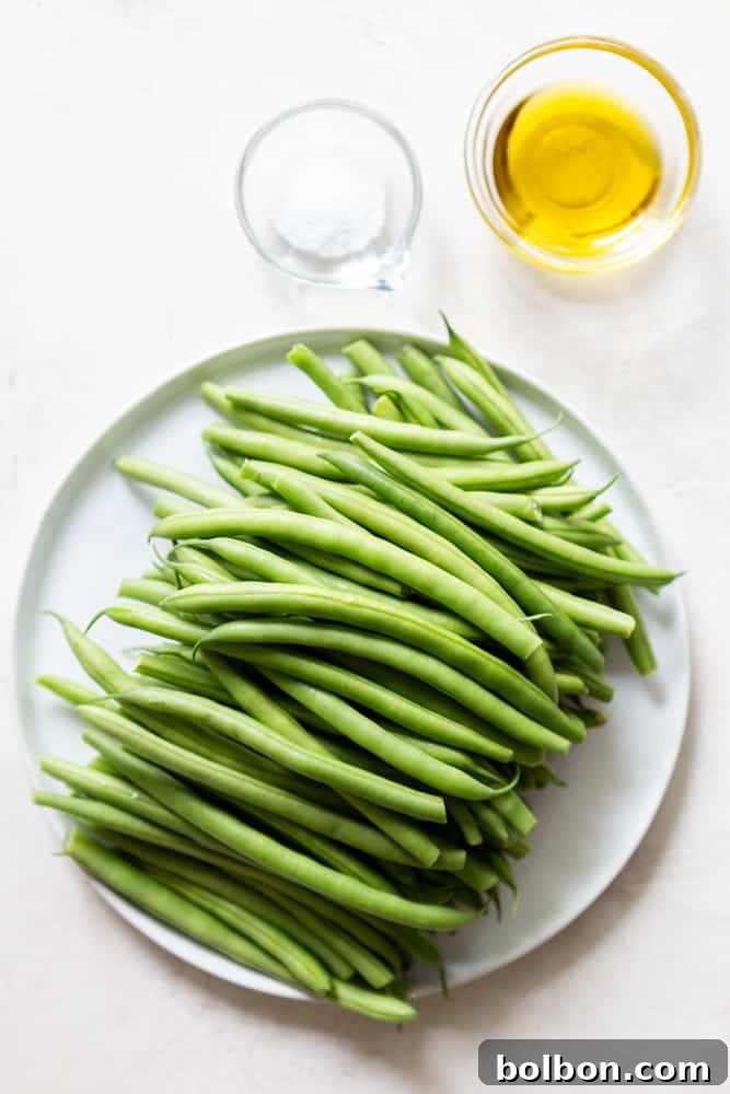 Fresh green beans arranged neatly on a white plate, accompanied by a small bowl of olive oil and a small bowl of salt, all set against a clean white background. This image highlights the fresh, simple ingredients required for the dish.