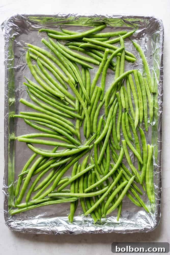 Uncooked fresh green beans spread evenly on a sheet pan, ready for roasting. The beans are vibrantly green and appear fresh, indicating good preparation before cooking.