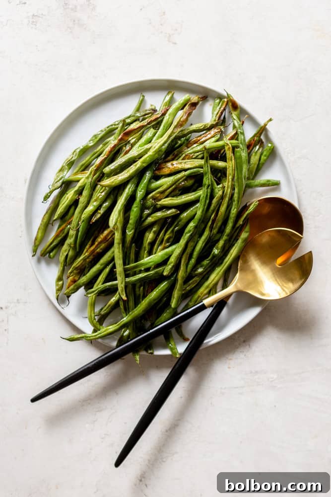 Oven roasted green beans presented on a white plate with elegant gold and black serving utensils. The dish looks appetizing and ready to be served.