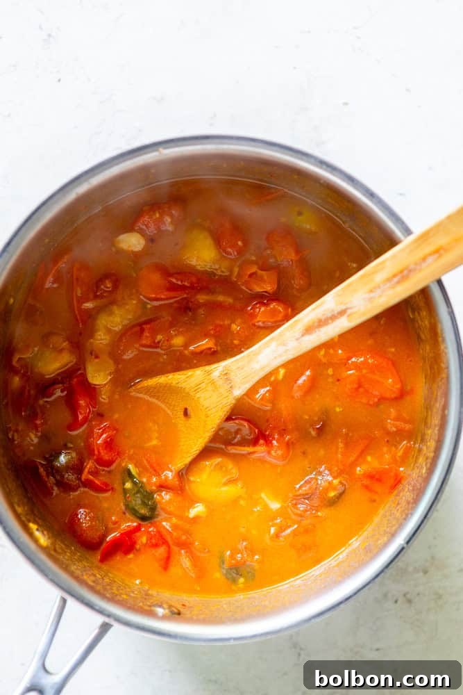 A close-up view of Burst Cherry Tomato Sauce simmering in a pan with a wooden spoon, showcasing the rich red color and tender, bursting tomatoes.