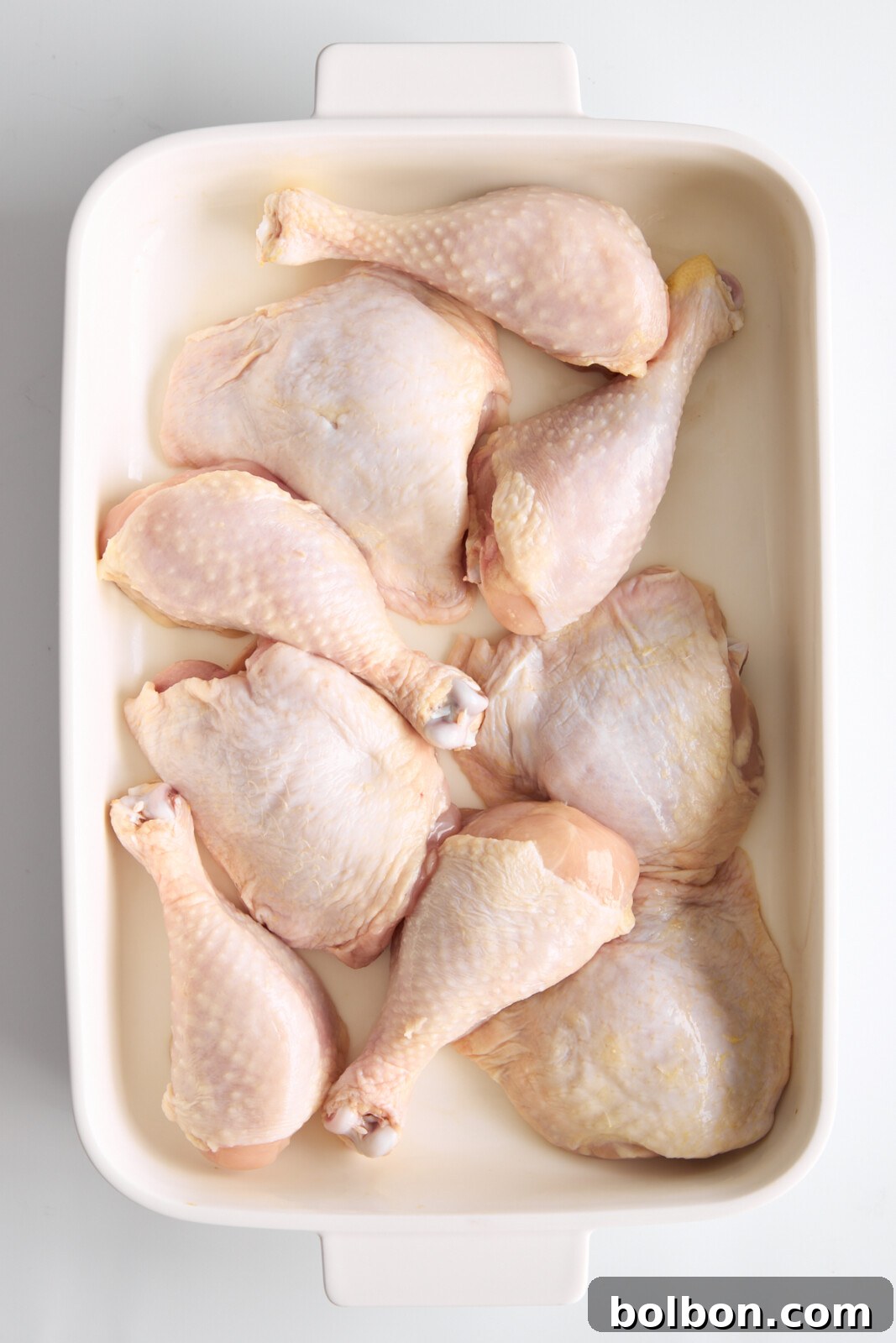 Zesty Greek Lemon Chicken 2 A white rectangular baking dish contains raw chicken drumsticks and thighs, neatly arranged. The chicken pieces have a light pink hue and delicate skin texture, indicating they are uncooked and ready for seasoning or cooking.