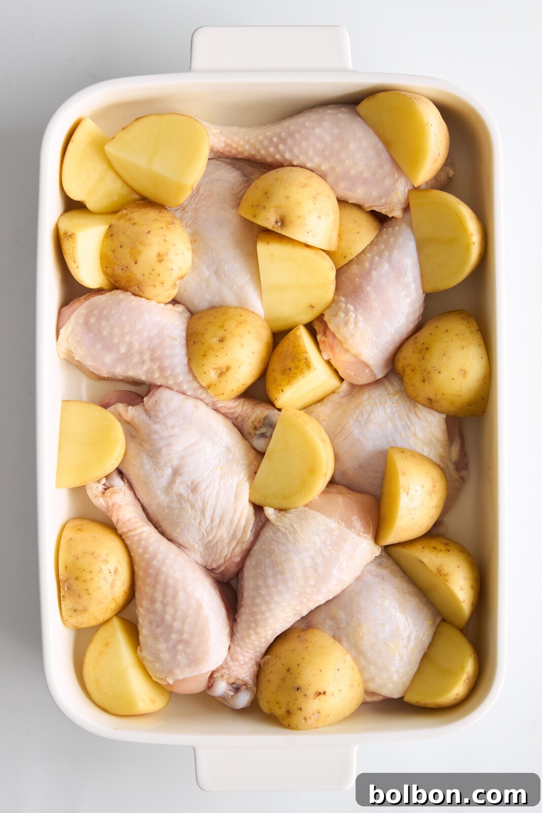 Zesty Greek Lemon Chicken 3 Top-down view of a white rectangular baking dish containing chicken drumsticks and halved yellow potatoes, prepared for cooking. The dish is set against a plain white background.