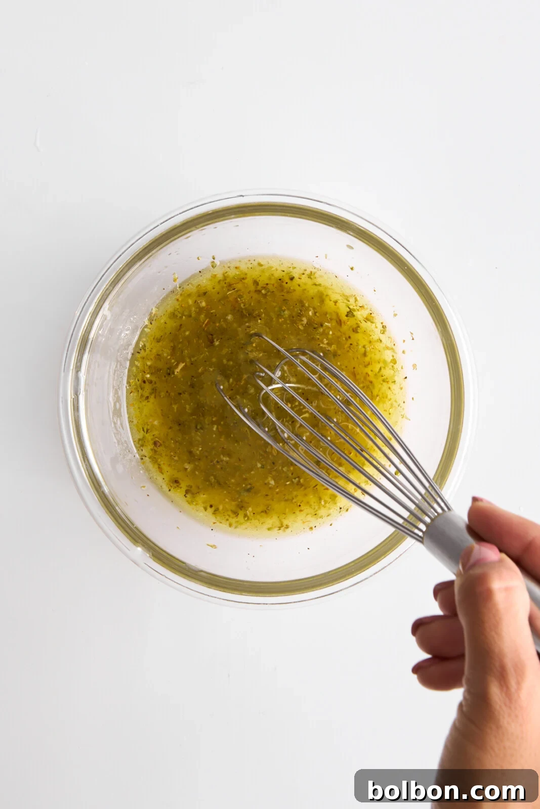 Zesty Greek Lemon Chicken 4 A hand whisking a yellow-green marinade or dressing in a clear glass bowl. The liquid contains visible herbs and spices, and the background is a plain white surface.