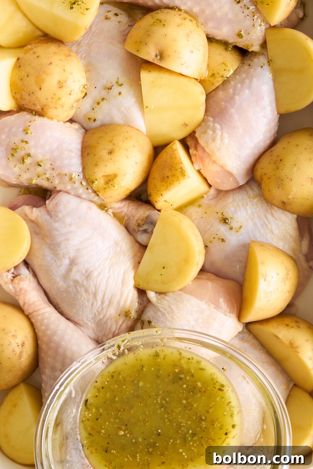 Zesty Greek Lemon Chicken 5 Raw drumsticks and halved baby potatoes are arranged in a roasting pan. A glass bowl with a mixture of olive oil and herbs is placed on top of some of the ingredients. The dish appears ready for baking.