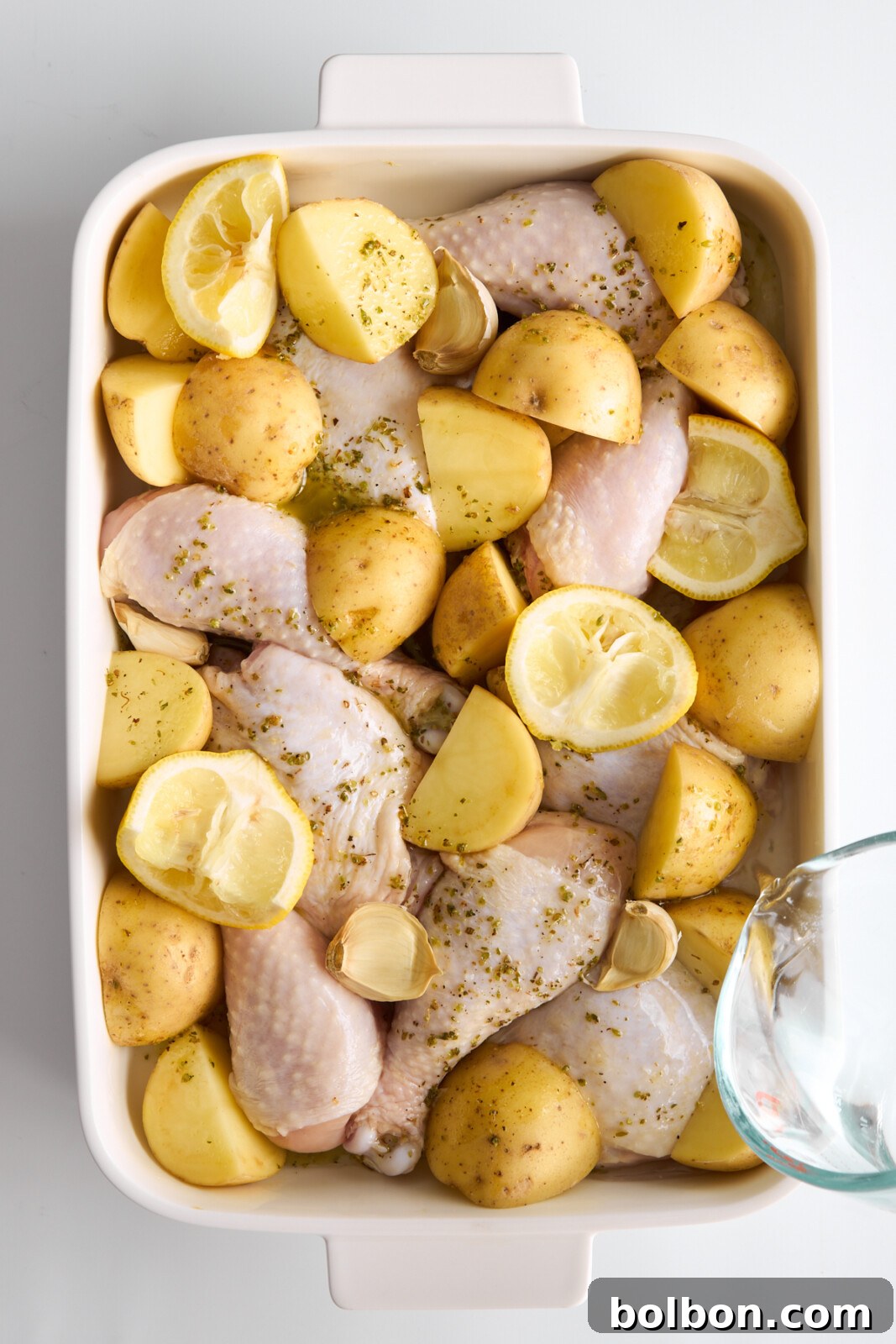 Zesty Greek Lemon Chicken 6 Greek Lemon Chicken ready to be baked. A white baking dish filled with raw drumsticks, potato wedges, garlic cloves, and sprinkled seasonings. The dish is prepared for baking, with a glass measuring cup pouring liquid into the corner of the dish.