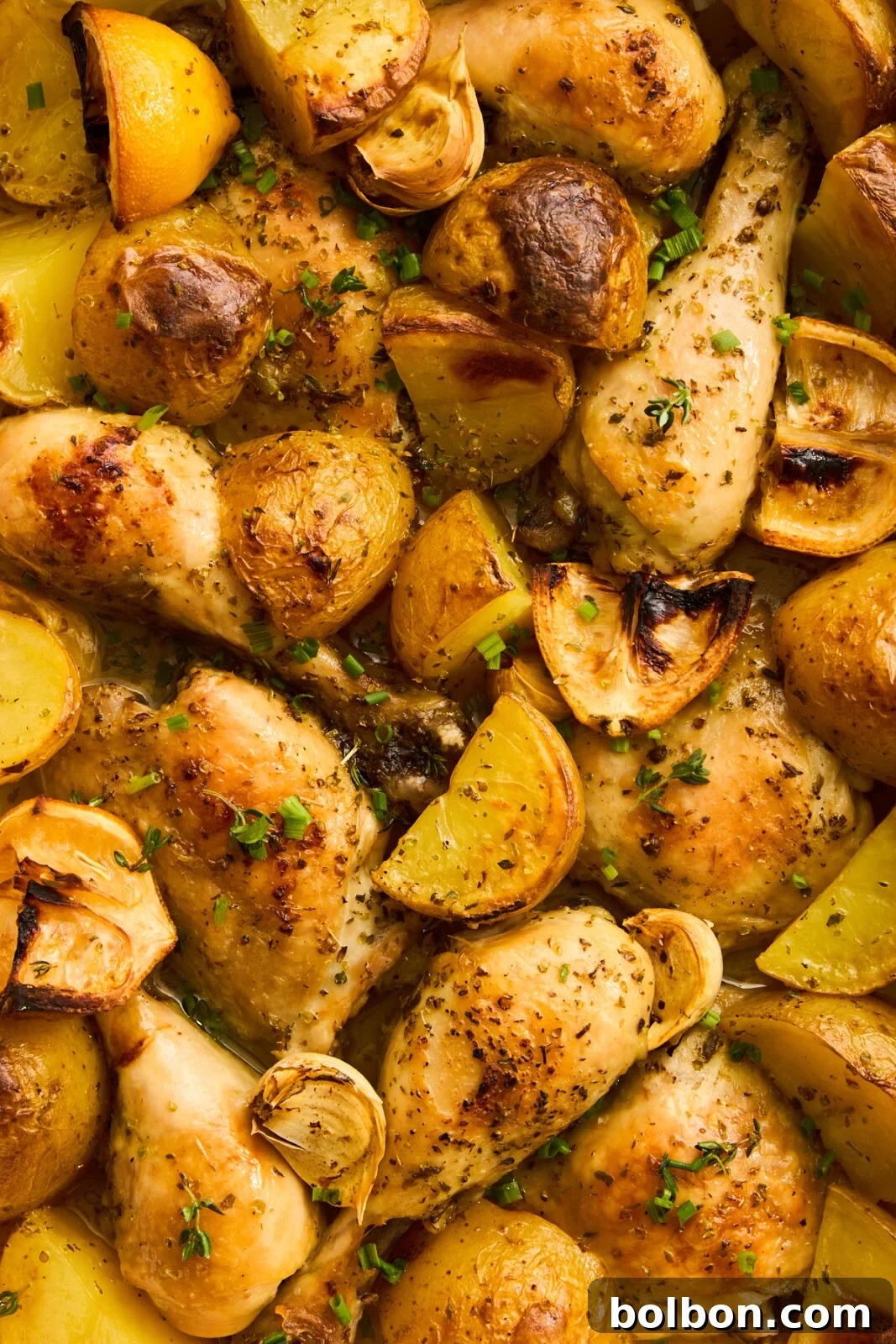 Zesty Greek Lemon Chicken 7 A close-up of a baked dish featuring seasoned chicken drumsticks and golden roasted potato wedges. The meal is garnished with fresh herbs, including chopped chives and parsley. The ingredients are evenly distributed, showcasing a crispy, well-cooked texture. Our Greek Lemon Chicken is ready.